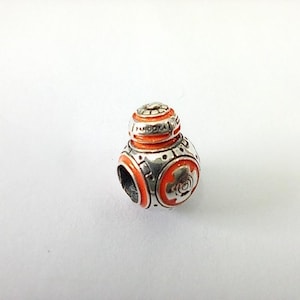 May include: A silver charm bead shaped like a spherical robot with orange enamel detailing. The metallic surface features engraved mechanical patterns and circular accents, with the brand name Pandora clearly visible on the upper section.