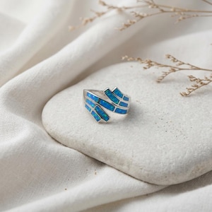 May include: A silver ring with a unique design featuring rectangular blue opal inlays. The ring is set on a white stone, with a soft white fabric in the background. The ring's design wraps around the finger, creating a modern and stylish look.