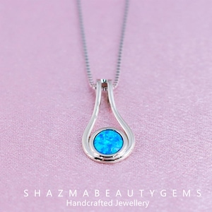 May include: A silver pendant necklace with a teardrop design. The pendant features a round, bright blue opal gemstone. The necklace is displayed on a pink background. The text "SHAZMABEAUTYGEMS Handcrafted Jewellery" is at the bottom.