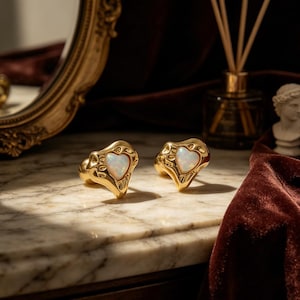 Puede incluir: Dos anillos dorados con una piedra de ópalo en forma de corazón. Los anillos tienen un diseño texturizado y se exhiben sobre una superficie de mármol. Los anillos son una joya.