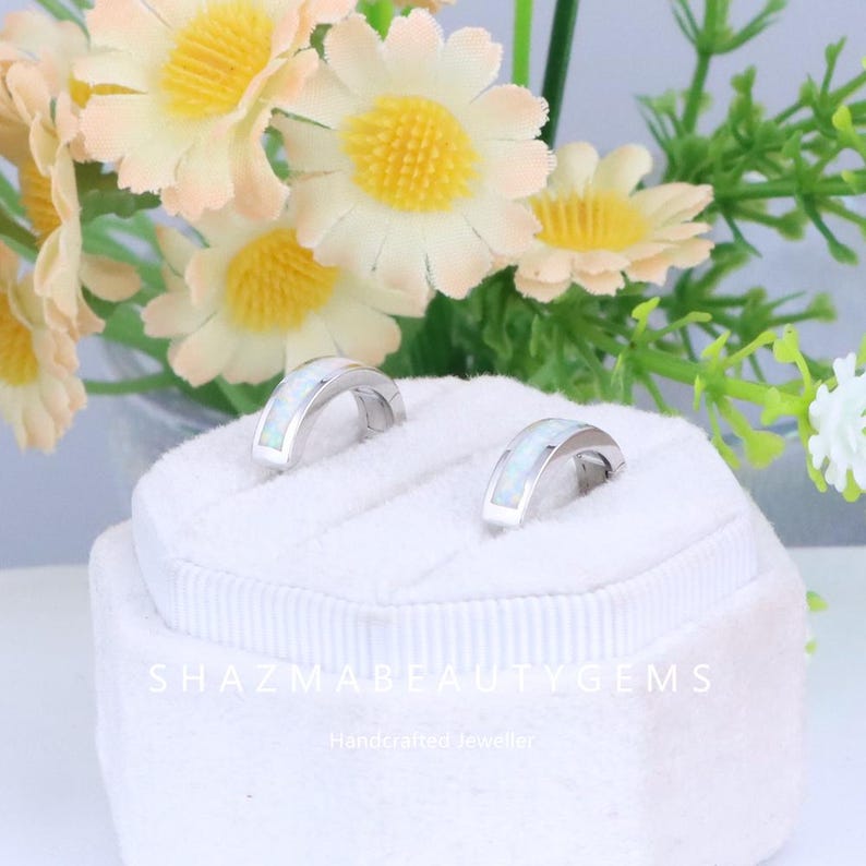 May include: Two silver-toned hoop earrings with a white opal inlay, displayed on a white velvet jewelry box. The earrings are set against a backdrop of artificial flowers with yellow and peach petals. The text "SHAZMABEAUTYGEMS" is visible.