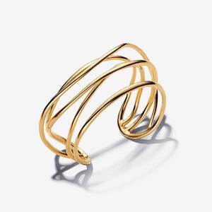 May include: A gold-toned cuff bracelet featuring a multi-strand design with four distinct, smooth, curved metal bands that overlap and intersect to create an open, airy structure with a polished, reflective finish and a wide, rounded opening.