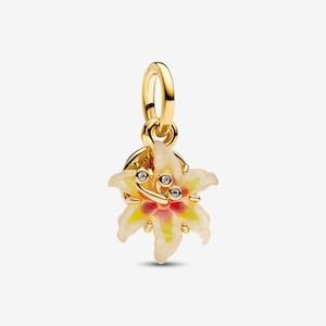 Puede incluir: Un dije dorado con forma de flor de lirio, que presenta pétalos de color crema con un centro rosa y amarillo. La flor está decorada con tres pequeñas piedras redondas y transparentes en su núcleo y cuelga de un anillo dorado simple y pulido.