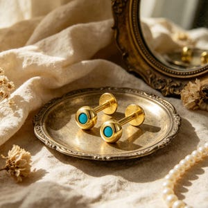 May include: Two gold-coloured cufflinks with turquoise blue centres, presented on a decorative silver-coloured plate. The cufflinks are round with a post and a flat back. A vintage mirror and a string of pearls are in the background.