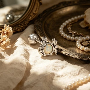 Puede incluir: Pendiente plateado con una piedra de ópalo redonda y detalles decorativos. El pendiente se muestra sobre una tela color crema, con un espejo de estilo vintage y joyas en el fondo.