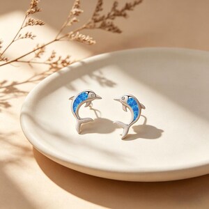 Peut inclure: Une paire de boucles d'oreilles dauphins en argent avec incrustation d'opale bleue et une petite gemme transparente. Les boucles d'oreilles sont présentées sur une assiette blanche avec un fond neutre. Accessoire fantaisiste.