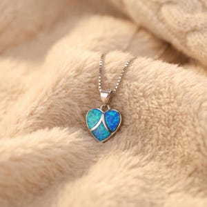 May include: Silver-toned necklace with a heart-shaped pendant. The pendant features a blue and teal opal design with a silver accent. The necklace is displayed on a soft, beige surface, showcasing the jewelry's delicate details.