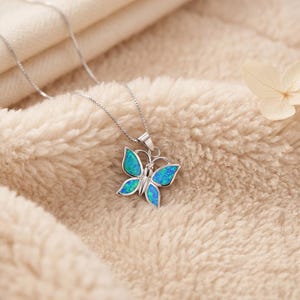 May include: A silver butterfly pendant necklace with a blue and green opal design. The butterfly pendant is detailed with a delicate silver outline and hangs from a fine silver chain. The necklace is displayed on a soft, cream-coloured surface.