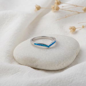 May include: A silver V-shaped ring with a band of blue opal stones. The ring is set on a white stone against a white fabric background. The ring is a piece of jewelry.