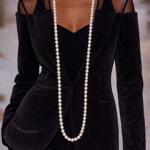 May include: A long strand of white pearls worn over a black velvet blazer with a V-neckline and sheer shoulder panels. The blazer features a single button closure, notched lapels, and long sleeves, creating a layered look with a black top underneath.