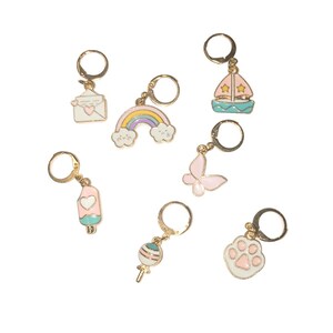 Op de afbeelding: Een verzameling goudkleurige oorringen met diverse pastelkleurige bedels. Bedels zijn onder meer een regenboog, vlinder, zeilboot, ijsje, pootafdruk, lolly en een envelop met een hart.