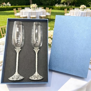 May include: A set of two clear glass champagne flutes with textured silver-toned metal stems and bases, presented in a dark blue rectangular gift box with a matching lid. The flutes feature tall, slender bowls and intricate, ornate metal detailing.