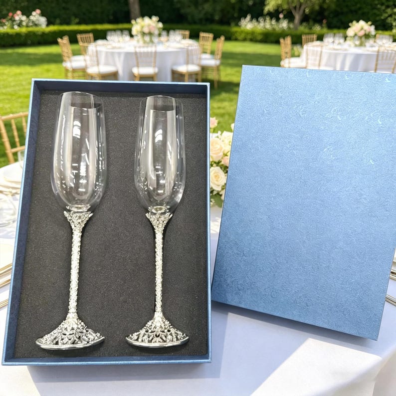 May include: A set of two clear glass champagne flutes with textured silver-toned metal stems and bases, presented in a dark blue rectangular gift box with a matching lid. The flutes feature tall, slender bowls and intricate, ornate metal detailing.