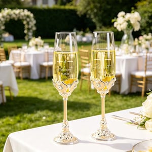 Peut inclure: Deux flûtes à champagne remplies d'un liquide doré, probablement du champagne, sont posées sur une surface blanche. Les flûtes ont des bases dorées ornées et sont décorées de bijoux étincelants. Chaque verre est gravé d'un nom et d'une date. Le fond suggère un mariage en plein air.