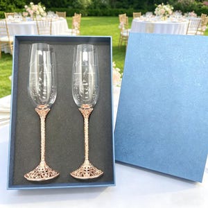 May include: Two clear glass champagne flutes with rose gold textured stems and bases, presented in a blue gift box. The glasses feature white etched text reading "Mark & Robert" with a date below. The stems have a detailed, intricate design.
