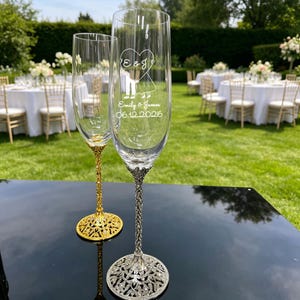 May include: Two clear glass champagne flutes with tall, slender bowls and ornate, textured stems. One stem is gold-toned, and the other is silver-toned. The front flute features an etched heart, the initials E & J, the names Emily & James, and 06.12.2026.
