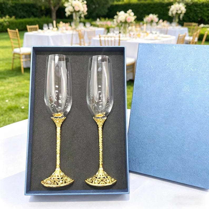 May include: A set of two clear glass champagne flutes with textured gold-toned metal stems and bases, presented in a blue rectangular gift box with a black foam insert. The flutes feature tall, slender bowls and intricate, ornate metal detailing.