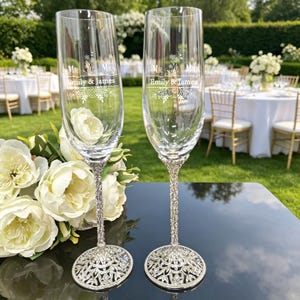 May include: Two clear glass champagne flutes with silver-toned, ornate, crystal-embellished stems and bases. The glass surface features white etched text reading "MR. & MRS." above "Emily & James" and a floral graphic design.
