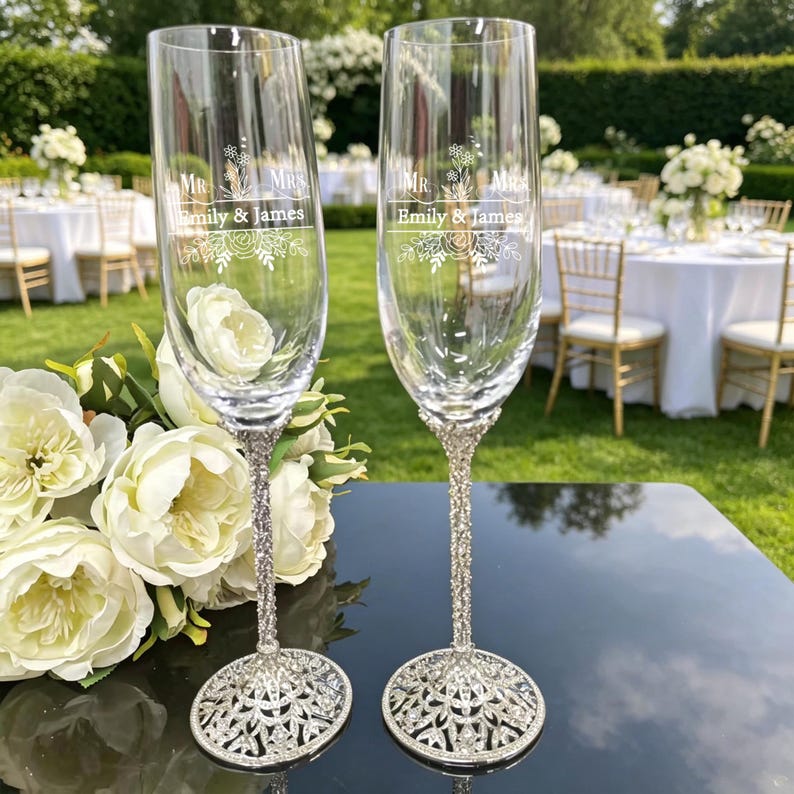 May include: Two clear glass champagne flutes with silver-toned, ornate, crystal-embellished stems and bases. The glass surface features white etched text reading "MR. & MRS." above "Emily & James" and a floral graphic design.