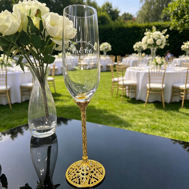 May include: A clear glass champagne flute with a gold-toned, ornate, filigree-style stem and base. The glass features white etched text that reads "we decided on forever", "Mr & Mrs", "Emily & James", and a date, with decorative flourishes.
