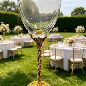 May include: A clear glass goblet featuring an ornate, textured gold-tone stem and base with intricate filigree detailing. The stem connects to the bowl with a decorative floral-inspired metal design, set against an outdoor event background.