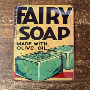 May include: Vintage metal sign for Fairy Soap, with a yellow background, black text, and two green soap bars. The sign reads "FAIRY SOAP MADE WITH OLIVE OIL". The sign has a distressed, aged appearance, suggesting it is an antique.