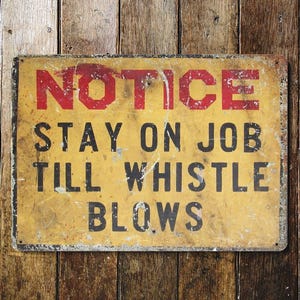 May include: A rectangular, vintage-style sign with the words "NOTICE STAY ON JOB TILL WHISTLE BLOWS" in red and black lettering. The sign has a yellow background with a distressed appearance, mounted on a wooden plank background.