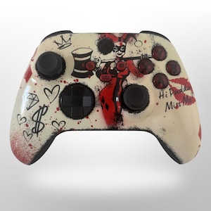 May include: Custom-designed video game controller with a cream-colored base. The controller features a Harley Quinn graphic, with red and black accents, and the words "Hi Puddin' Miss Me". Additional graphics include hearts, diamonds, and a crown.