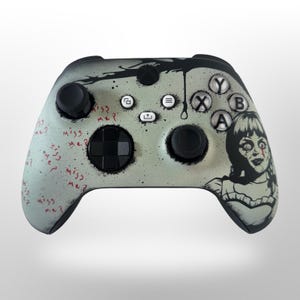 May include: Custom-painted video game controller with a horror theme. The controller has a light green and gray speckled background with a black design at the top. The controller features a black and white illustration of a doll with the words "miss me?" in red.