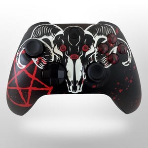 May include: Black Xbox controller featuring a white ram skull design and red accents. The controller has a pentagram on the left and red splatter details. The buttons are black and red.