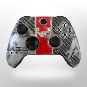 Puede incluir: Un mando de videojuegos con patrón de fibra de carbono y acabado plateado y rojo desgastado. El diseño incluye el texto Mass Effect, una franja roja central, joysticks negros, una cruceta negra y botones de acción blancos etiquetados Y, X, B, A.