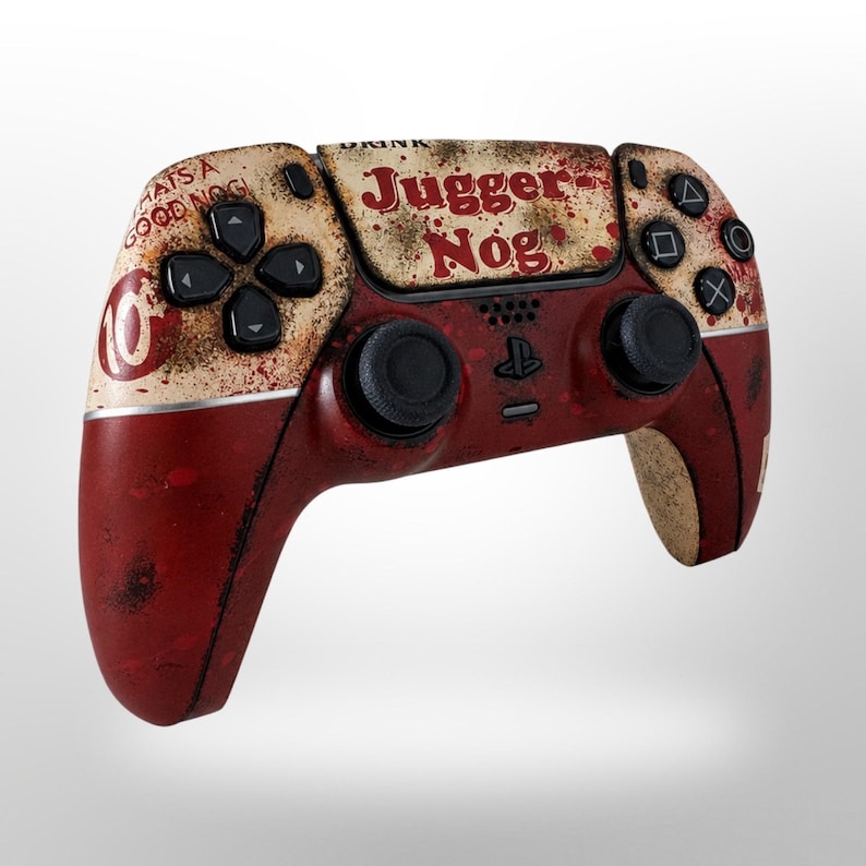 May include: A custom PlayStation 5 controller with a distressed, vintage design. The controller features a red and cream color scheme with the words "Juggernog" and "Whats a Good Nog" printed on it. The buttons and joysticks are black.