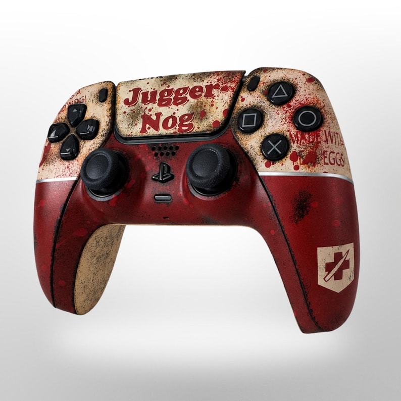 May include: A custom PlayStation 5 controller with a distressed, weathered design. The controller is primarily red with beige accents and blood splatter details. The text "JUGGER NOG" is displayed on the top, with "MADE WITH EGGS" on the side.