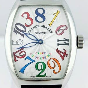 Puede incluir: Reloj de pulsera plateado con esfera blanca y grandes números coloridos. La esfera muestra las palabras "FRANCK MULLER GENEVE" y "CRAZY COLOR DREAMS". Tiene una correa de cuero negro y una corona texturizada.