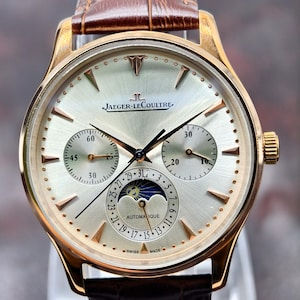 Può includere: Un orologio automatico Jaeger-LeCoultre color oro con quadrante argentato a raggiera, dotato di due sotto-quadranti, una fase lunare e un indicatore della data. L'orologio presenta indici e lancette color oro e un cinturino in pelle marrone.