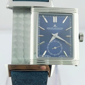 Könnte beinhalten: Eine rechteckige silberne Armbanduhr mit blauem Zifferblatt und blauem Lederarmband. Das Zifferblatt hat silberne Zeiger und Markierungen mit dem Markennamen "JAEGER-LECOULTRE". Sie hat eine kleine Sekundenscheibe und eine Krone.