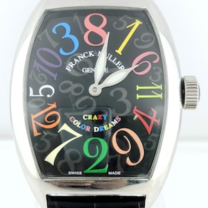 Könnte beinhalten: Eine silberfarbene Franck Muller Crazy Color Dreams Uhr mit schwarzem Zifferblatt. Die Uhr hat farbige Ziffern und die Worte "CRAZY COLOR DREAMS". Die Uhr hat ein schwarzes Lederarmband.