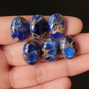 Puede incluir: Seis piedras preciosas ovaladas de color azul intenso con vetas doradas. Las piedras pulidas se sostienen en una mano, mostrando sus superficies reflectantes y patrones únicos. Ideales para la creación de joyas.