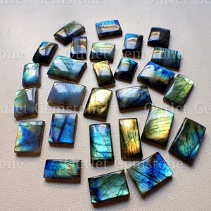 May include: A collection of rectangular labradorite gemstones featuring iridescent flashes of blue, gold, and green. Each polished stone displays unique internal fractures and colour shifts across its surface, arranged in a scattered cluster.