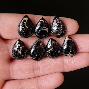 May include: Eight black teardrop-shaped gemstones with gold veining are displayed. The polished stones have a glossy finish. The gemstones are held in a hand against a dark background. Each stone is approximately 2 cm in length.