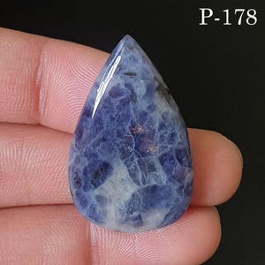 Puede incluir: Una gema pulida en forma de lágrima. La piedra presenta una mezcla de tonos azul oscuro y blanco, con sutiles motas doradas. La gema se sostiene entre los dedos, con el texto "P-178" en la esquina superior derecha.