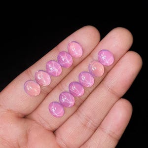 Puede incluir: Diez piedras preciosas rosas de forma ovalada dispuestas en dos filas, cada una con un patrón de estrella de seis puntas conocido como asterismo. Estas piedras cabujón lisas y pulidas exhiben una calidad translúcida en tonos rosados.
