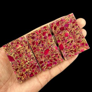 Può includere: Tre piastrelle rettangolari a mosaico caratterizzate da un mix vibrante di frammenti di pietra magenta scuro e rosa brillante incastonati in una matrice metallica color oro scintillante, creando una superficie densa e riflettente.