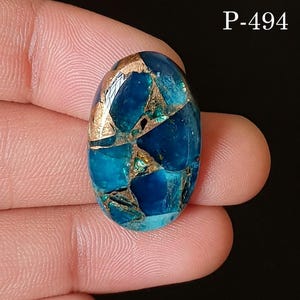 Peut inclure: Pierre précieuse ovale avec une mosaïque de fragments bleu foncé et turquoise, entrecoupés de veines métalliques dorées. La surface polie est réfléchissante. Le texte "P-494" est visible en haut à droite.