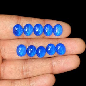 Puede incluir: Diez piedras preciosas cabujón de color azul dispuestas en dos filas. Cada piedra presenta un patrón de estrella de seis puntas, conocido como asterismo, visible en la superficie lisa y pulida. Las piedras exhiben un color azul vibrante.