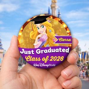 Op de afbeelding: Een ronde, paarse en gouden afstudeerknop met een cartoon van een vrouw met lang blond haar en een afstudeerhoed. De tekst luidt "Just Graduated Class of 2026" en "Cierra". Het Walt Disney World-logo staat onderaan.