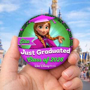 May include: A round, purple graduation button featuring the character Anna from Disney's Frozen. The button reads "Ella Just Graduated Class of 2026" with the Walt Disney World logo. The background shows a blurred theme park.