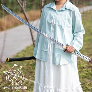 Könnte beinhalten: Ein langes, silbernes Katana-Schwert mit schwarzem Griff und Scheide. Das Schwert wird von einer Person gehalten, die ein hellblaues Hemd und ein weißes Kleid trägt. Das Schwert hat einen dekorativen Griff und eine scharfe Klinge. Das Logo "SteelHarmonyCrafts" ist sichtbar.