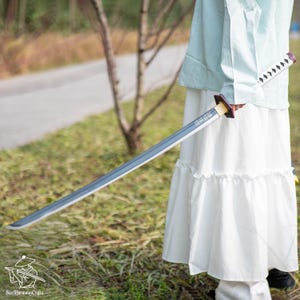 May include: A katana sword with a gray blade and a white handle with black accents. The hilt is brown and gold. The sword is held by a person wearing a white skirt and a light blue top. The sword has Japanese text on the blade.