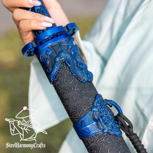 May include: Close-up of a blue and black decorative sword. The sword features intricate dragon designs in blue on the hilt and scabbard. The scabbard is black with a detailed pattern. The sword is held by a person wearing a light blue garment. The logo "SteelHarmonyCrafts" is visible.